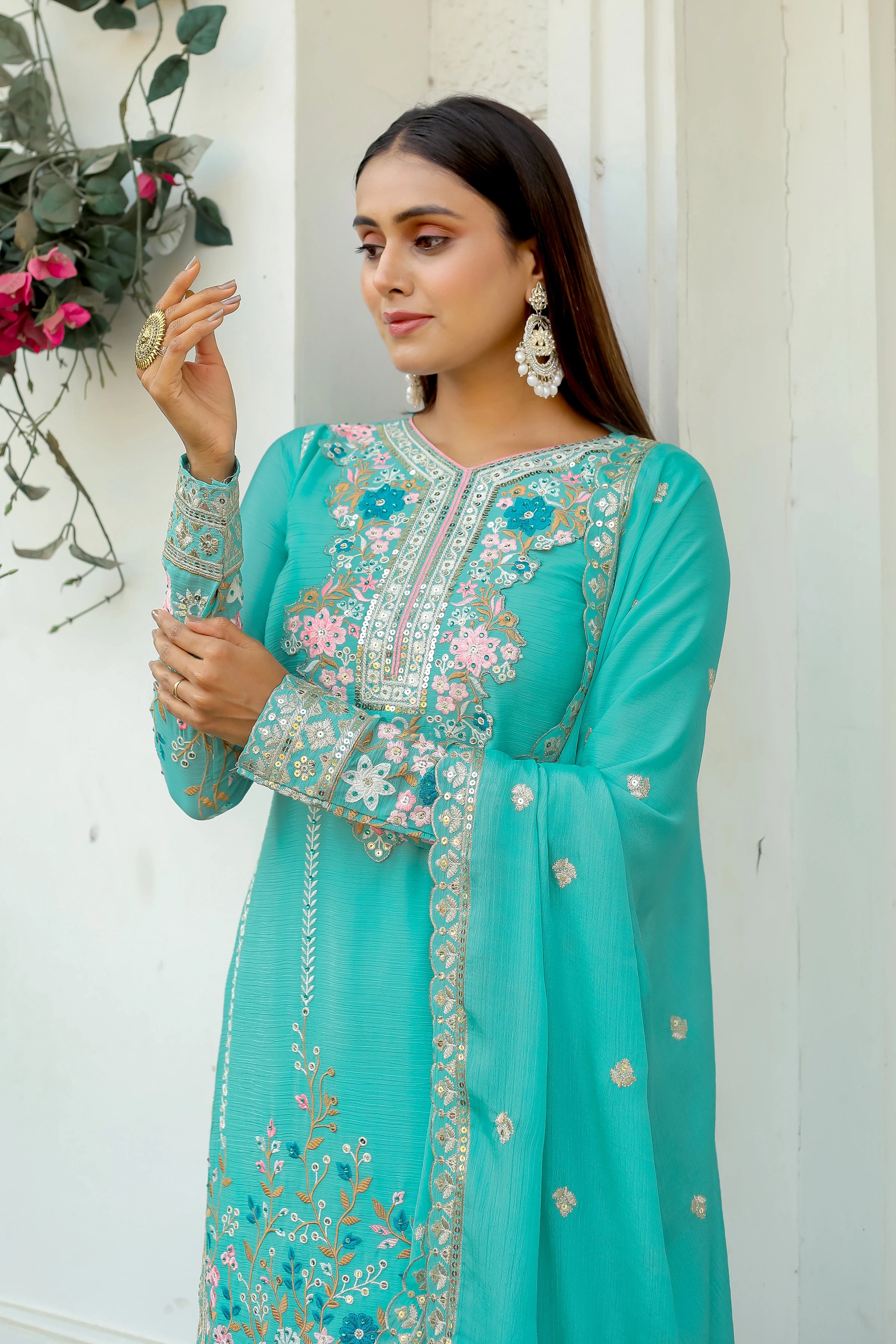 Woman wearing a turquoise embroidered outfit holding a clutch against a white wall.